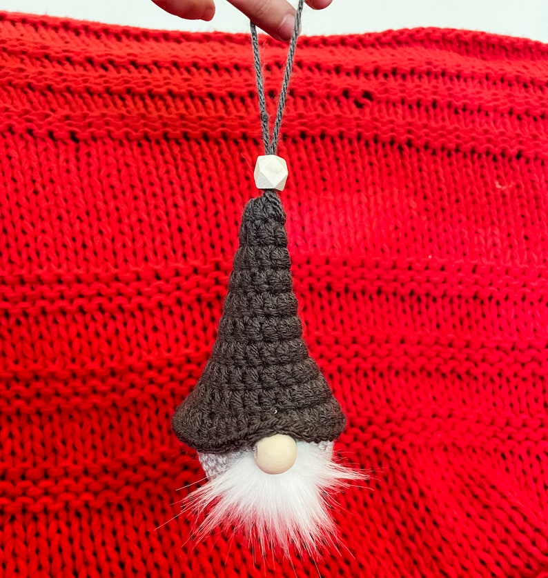 May include: A black and white gnome ornament with a white beard and a wooden bead nose. The gnome is hanging from a red knitted background.