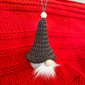 May include: A crocheted gnome ornament with a black hat and white beard. The gnome is hanging from a red knitted background.