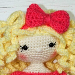 Pu&ograve; includere: Una bambola all'uncinetto fatta a mano con un viso color crema, occhi di perline nere e una piccola bocca rosa. La bambola ha capelli ricci gialli e un fiocco rosa brillante sulla testa. La bambola indossa un vestito rosa.