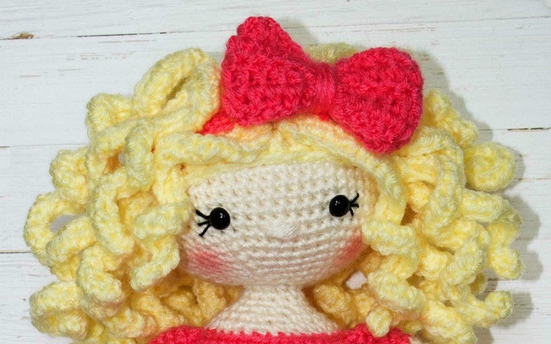 Pu&ograve; includere: Una bambola all'uncinetto fatta a mano con un viso color crema, occhi di perline nere e una piccola bocca rosa. La bambola ha capelli ricci gialli e un fiocco rosa brillante sulla testa. La bambola indossa un vestito rosa.
