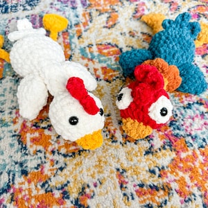 May include: Two crocheted stuffed chickens, one white with yellow beak and feet, the other blue with red and orange accents and a yellow beak.