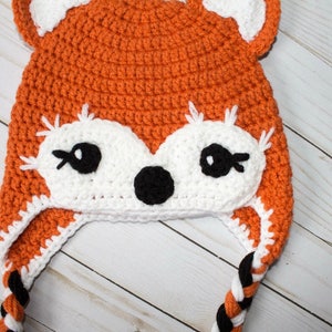 Puede incluir: Sombrero de zorro de crochet naranja y blanco con lazos trenzados negros. El sombrero tiene una cara blanca con ojos negros y una nariz negra.