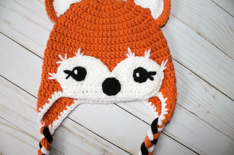 Puede incluir: Sombrero de zorro de crochet naranja y blanco con lazos trenzados negros. El sombrero tiene una cara blanca con ojos negros y una nariz negra.