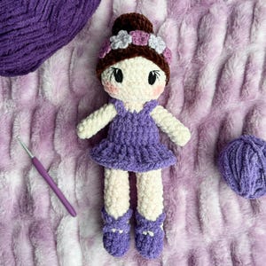 May include: A crocheted doll with a purple dress and a flower crown. The doll has brown hair and white skin. The doll is sitting on a purple and white fuzzy blanket.