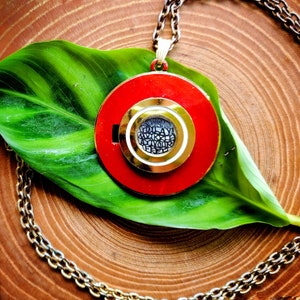 Op de afbeelding: Een rode en gouden ketting met een ronde hanger met een cirkelvormig ontwerp met zwarte tekst. De hanger ligt op een groen blad.