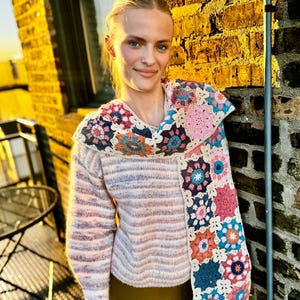 May include: A light pink and white striped sweater with a colorful crochet scarf featuring floral patterns in shades of pink, blue, and orange. The scarf has a fringed edge. The person is wearing a dark green skirt.