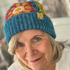 Op de afbeelding: Een gehaakte muts met een kleurrijk granny square patroon in rood, geel en groen op de kruin, met een effen blauwgroene geribbelde rand. Een klein rechthoekig beige leren label met de tekst "Chro gola 57" is aan de zijkant bevestigd.