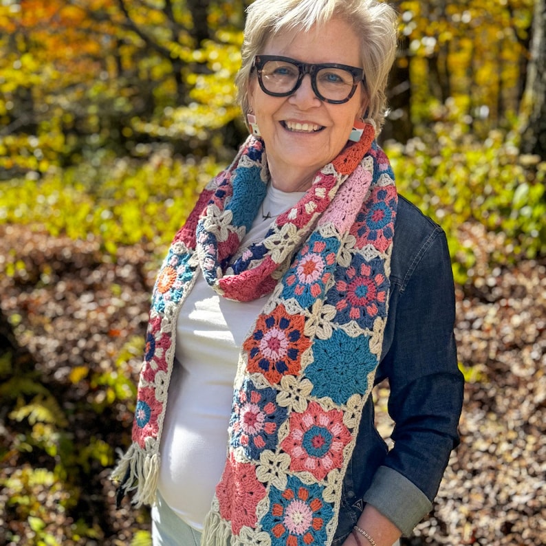 May include: A colorful, crocheted scarf with a patchwork design. The scarf features squares in shades of pink, blue, orange, and cream, with a floral pattern. The scarf has fringe at the ends and is worn around the neck.