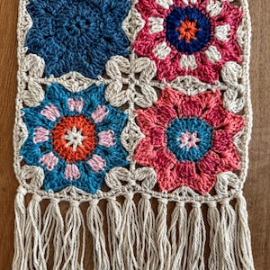 May include: A handmade crochet table runner featuring a repeating floral pattern in shades of blue, pink, orange, and cream. The runner has a cream-colored border and fringe along the bottom edge. The floral pattern is made of squares.