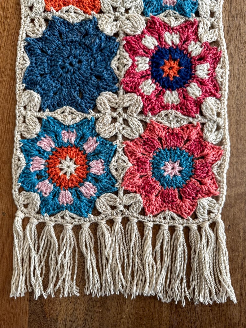 May include: A handmade crochet table runner featuring a repeating floral pattern in shades of blue, pink, orange, and cream. The runner has a cream-colored border and fringe along the bottom edge. The floral pattern is made of squares.