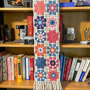 May include: A handmade crochet scarf with a floral pattern in shades of coral, blue, and orange, connected by a cream-colored border. The scarf has a fringe at the bottom and is displayed on a wooden hanger.