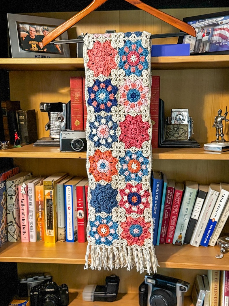 May include: A handmade crochet scarf with a floral pattern in shades of coral, blue, and orange, connected by a cream-colored border. The scarf has a fringe at the bottom and is displayed on a wooden hanger.