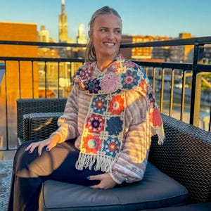 May include: A colorful crochet scarf with a square pattern in shades of pink, blue, orange, and cream. The scarf has a fringed edge and is draped over a striped sweater. The background shows a city skyline at sunset.
