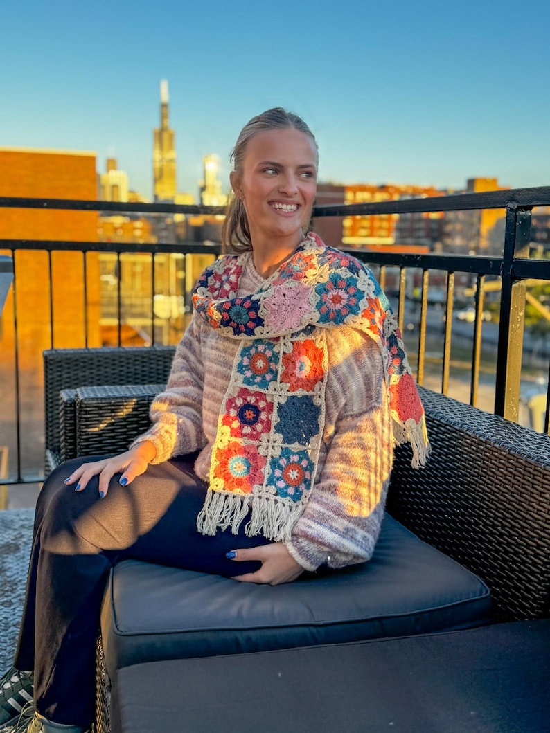 May include: A colorful crochet scarf with a square pattern in shades of pink, blue, orange, and cream. The scarf has a fringed edge and is draped over a striped sweater. The background shows a city skyline at sunset.