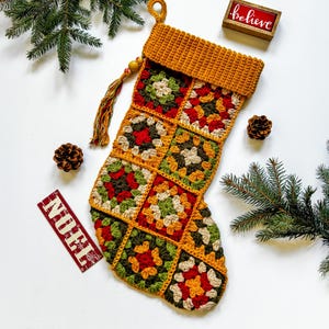 Könnte beinhalten: Ein handgefertigter Weihnachtsstrumpf mit einem Granny-Square-Design in Rot-, Grün-, Gelb- und Cremetönen. Der Strumpf hat eine senfgelbe Manschette und Schlaufe mit Quaste. Das Wort "believe" steht auf einem kleinen Holzschild.