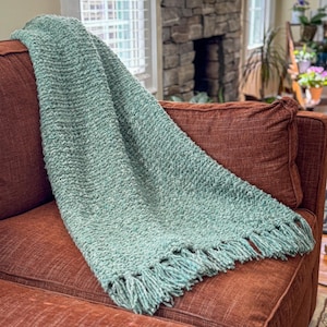 May include: A seafoam green throw blanket with a textured weave and fringed edge, draped over a rust-coloured sofa. The blanket's thick, cosy material suggests warmth and comfort, adding a stylish touch to a living room setting.