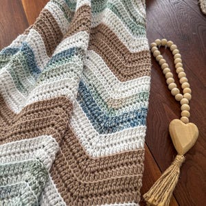 May include: A crocheted blanket with a chevron design in brown, white, blue, and green. The blanket is folded, displaying its texture and colour variations. A wooden heart-shaped ornament with a tassel hangs nearby.