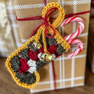 May include: A wrapped gift with brown paper and red twine, adorned with a crocheted Christmas stocking in yellow, green, red, and white. Two small gold bells and a candy cane complete the festive look.