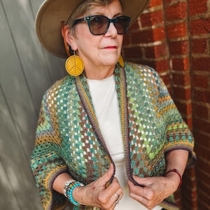 May include: A woman wearing a tan hat, sunglasses, and yellow peace sign earrings. She is wearing a white shirt and a colorful, open-front crochet cardigan in shades of green, yellow, and brown. The cardigan has a geometric pattern. She is also wearing bracelets.