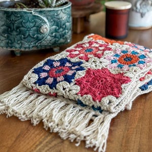 May include: A handmade crochet scarf featuring a colorful floral pattern in shades of red, blue, orange, and pink. The scarf has a neutral-colored base and is finished with a fringe trim. The scarf is displayed on a wooden surface.