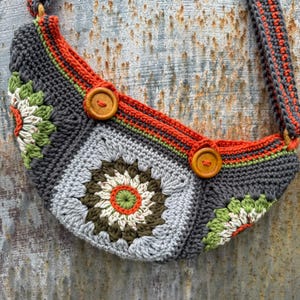 Może przedstawiać: Ręcznie robiona torba z szydełka z kwiatowym wzorem. Torba ma szarą podstawę z pomarańczowymi, zielonymi i białymi akcentami. Dwa drewniane guziki zabezpieczają klapę. Pasek i wykończenie są pomarańczowe i szare.