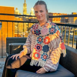 May include: A colorful, crocheted scarf with square floral patterns in shades of pink, blue, and orange, fringed at the ends. The scarf is draped over a sweater. The background shows a city skyline.