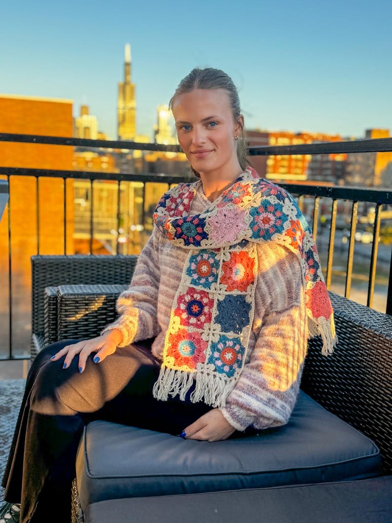 May include: A colorful, crocheted scarf with square floral patterns in shades of pink, blue, and orange, fringed at the ends. The scarf is draped over a sweater. The background shows a city skyline.