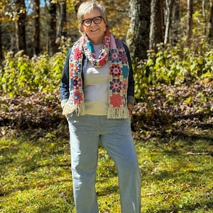 May include: A colorful crochet scarf with a floral pattern and fringe is worn over a denim jacket and a light-colored shirt. The person is wearing light blue pants and glasses, standing in a forest setting with green grass and trees.