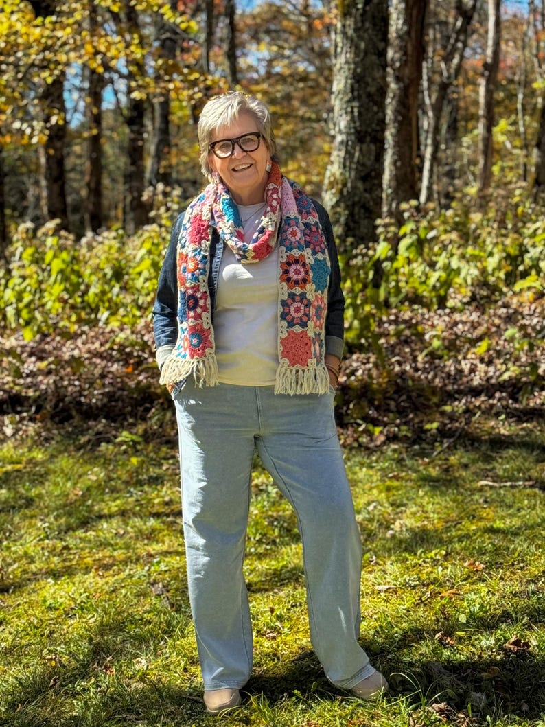 May include: A colorful crochet scarf with a floral pattern and fringe is worn over a denim jacket and a light-colored shirt. The person is wearing light blue pants and glasses, standing in a forest setting with green grass and trees.