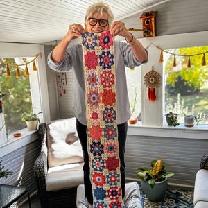 May include: A colorful, handmade crochet scarf featuring a floral pattern in shades of red, pink, blue, and orange. The scarf has a cream-colored background and a fringed edge. The scarf is held up in front of a window.