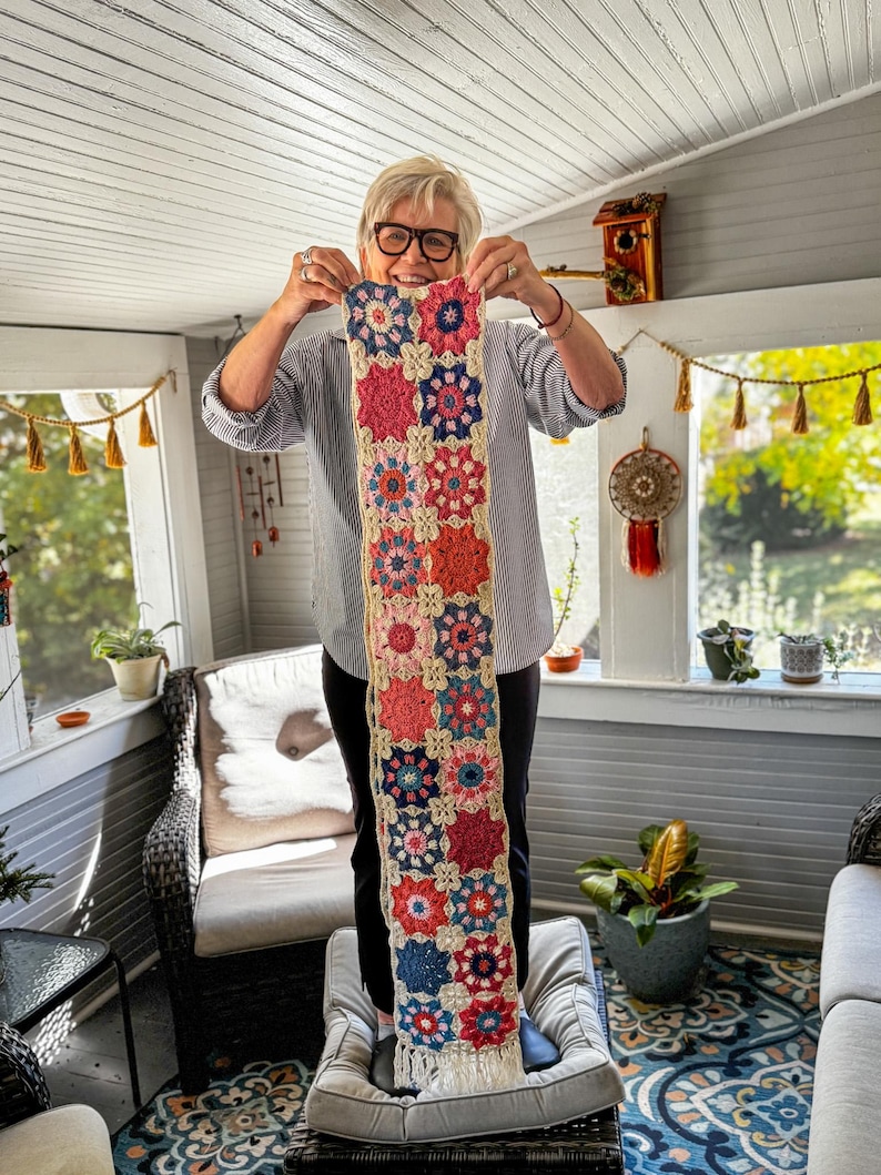 May include: A colorful, handmade crochet scarf featuring a floral pattern in shades of red, pink, blue, and orange. The scarf has a cream-colored background and a fringed edge. The scarf is held up in front of a window.