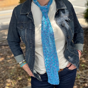 May include: A blue and purple crochet tie with a textured pattern. The tie is worn over a white shirt and a denim jacket. The tie's color gradient includes shades of light blue and purple.
