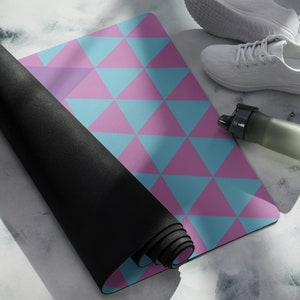 Puede incluir: Una esterilla de yoga negra enrollada con un patrón geométrico rosa y azul en la parte superior. La esterilla está parcialmente desenrollada, revelando el patrón. Una botella de agua blanca y un par de zapatillas blancas están en el fondo.