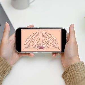 May include: A smartphone screen shows a divination chart with a semi-circular design and text. The chart is peach-coloured with dark lines and text. The phone is held in front of a white surface, with a laptop, mug, and plant in the background.