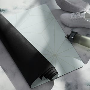 Puede incluir: Una esterilla de yoga negra enrollada con un patrón geométrico blanco y dorado en la parte superior. La esterilla está parcialmente desenrollada, revelando el patrón. También se ve una botella de agua blanca y unas zapatillas blancas en la imagen.
