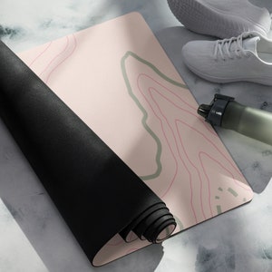 Puede incluir: Una esterilla de yoga enrollada con un diseño de mapa topográfico en rosa, verde y rojo. Un par de zapatillas blancas y una botella de agua están junto a la esterilla. La parte inferior de la esterilla es negra.