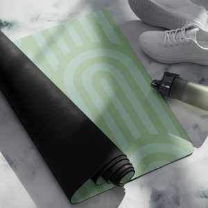 Puede incluir: Una esterilla de yoga verde y blanca enrollada con un patrón geométrico. La esterilla está sobre una superficie gris claro con una botella de agua blanca y zapatillas blancas.