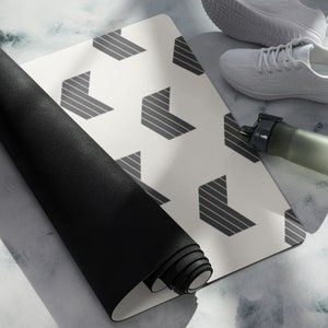 Puede incluir: Una esterilla de yoga blanca con un patrón geométrico de flechas en blanco y negro. La esterilla está enrollada y una esterilla de yoga negra está encima. Una zapatilla blanca y una botella de agua están en el fondo.