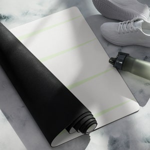 Puede incluir: Una esterilla de yoga negra enrollada con líneas verde claro sobre un fondo blanco. También se ve un zapato deportivo blanco, una botella de agua transparente y una superficie blanca.