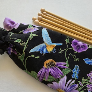 May include: A black drawstring knitting needle case with a floral and bird design. The case features a pattern of purple flowers, blue birds, and green vines. Several wooden knitting needles are next to the case.