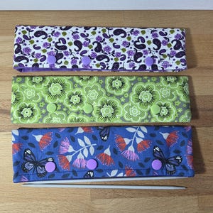 May include: Three fabric knitting needle cases with button closures. The top case is white with purple paisley, the middle is green with floral patterns, and the bottom is blue with butterflies and floral designs. A knitting needle is placed below the cases.