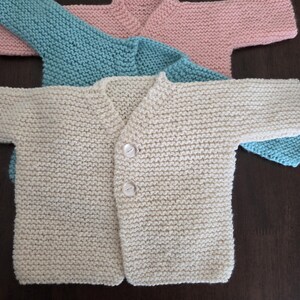 May include: Three hand-knitted baby cardigans in pastel shades: cream, light blue, and pink. The cream cardigan is fastened with two white buttons. The cardigans are made of a soft, textured yarn, ideal for a newborn.