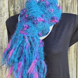 May include: A hand-knitted scarf in shades of turquoise, purple, and pink. The scarf has a textured, fluffy appearance with long fringe. The scarf is displayed on a black top against a weathered wooden background.