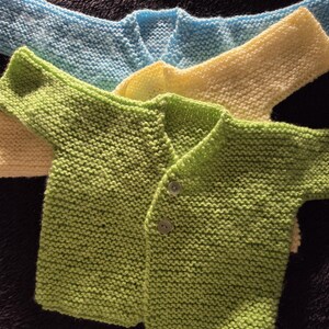 May include: Close-up of three hand-knitted baby cardigans in shades of green, yellow, and blue. The green cardigan in the foreground has two small buttons. The cardigans are made of a soft, textured yarn.