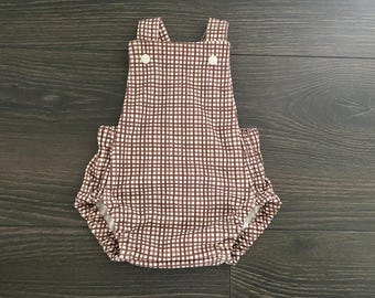May include: A brown and white gingham print baby romper featuring a square neckline, two shoulder straps with white buttons, and elasticized leg openings. The garment has a gathered design at the sides and a simple, sleeveless construction.