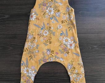 May include: A sleeveless mustard yellow romper featuring a floral pattern with white daisies, purple blossoms, and green foliage. The garment has a round neckline, two white snap buttons at the shoulders, and cuffed leg openings at the bottom.