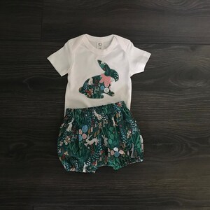 May include: A white bodysuit with a green and white bunny applique. The bodysuit is paired with green and white floral print bloomers.