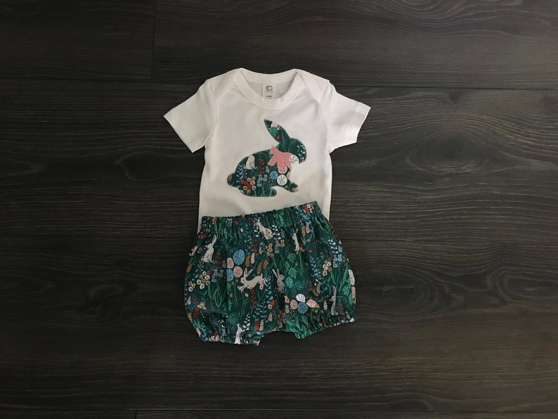 May include: A white bodysuit with a green and white bunny applique. The bodysuit is paired with green and white floral print bloomers.