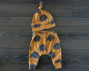 May include: A set of baby clothes featuring a brown and black patterned hat and matching pants with a print of echidnas. The hat has a knotted top and the pants have an elastic waistband.