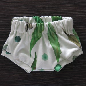 May include: White baby bloomers with an elastic waistband, featuring a watercolor print of green leaves and dots. The fabric is soft and the design is gender-neutral.